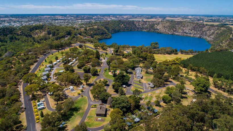 Aerial view of Mount Gambier showcasing the Blue Lake, surrounded by greenery, residential areas, and farmland under a clear sky.